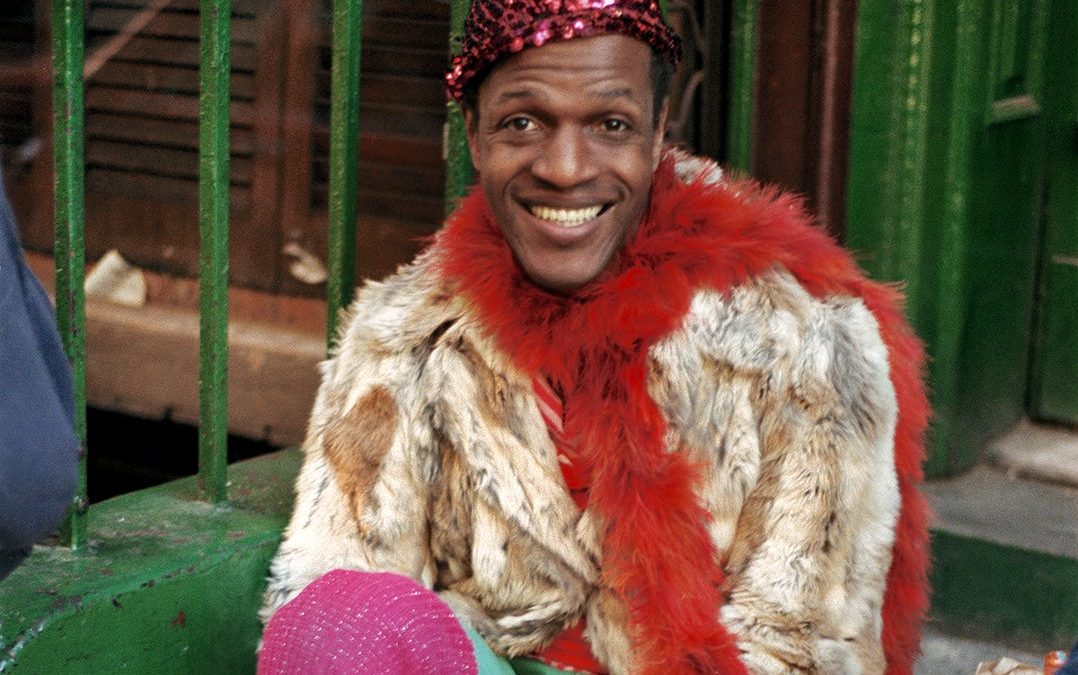 Marsha P Johnson seated on steps wearing a sequin pink hat, fur coat, red fur boa, and pink leg warmers smoking a cigarette.