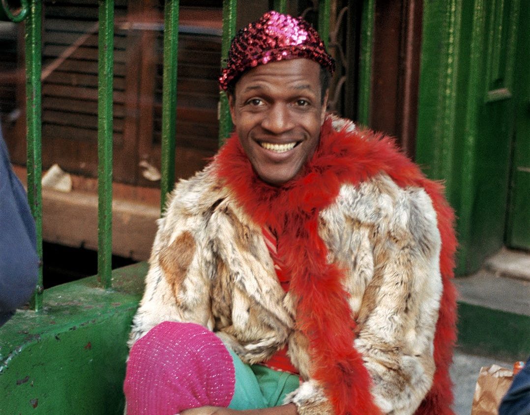 Marsha P Johnson seated on steps wearing a sequin pink hat, fur coat, red fur boa, and pink leg warmers smoking a cigarette.