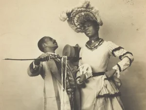 Black and white photograph of two Black men. One is dressed in male attire, including a top hat. The other is wearing a white ruffled dress and elaborate hat.