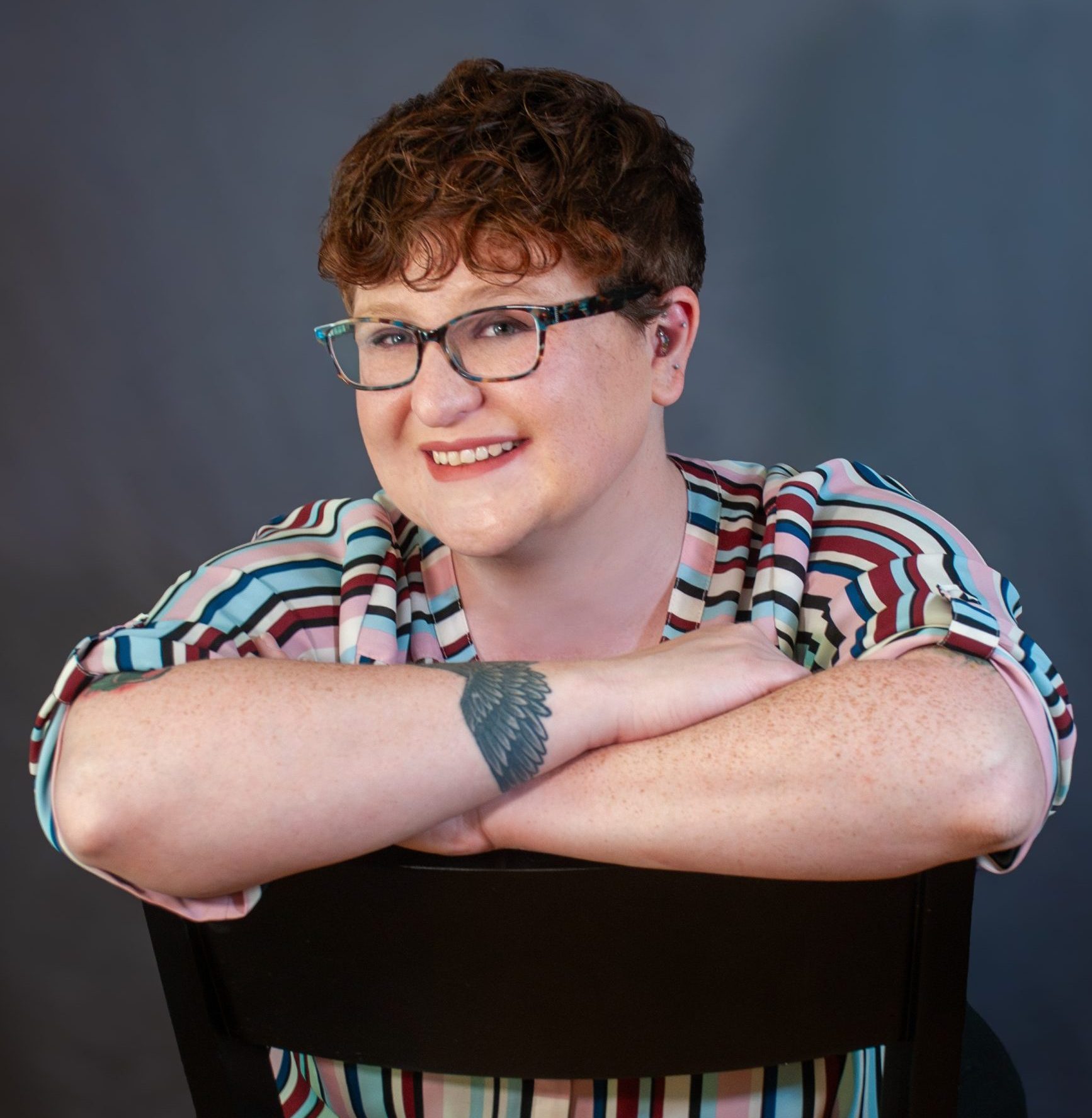 Ali sits backwards in a black chair, crossing her arms over the back of the chair. Ali is a pale-skinned individual with a short auburn hair and blue plastic frame glasses.