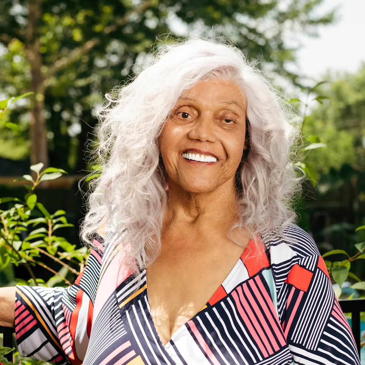 Miss Major, an elderly woman with tousled gray hair, smiles at the camera against lush greenery.