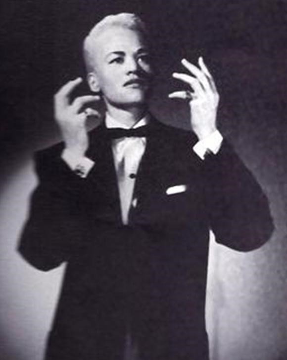 Black and white Portrait of Stormé DeLarverie, who has a short cropped haircut and is wearing a tuxedo. She is posing as if presenting a monologue.