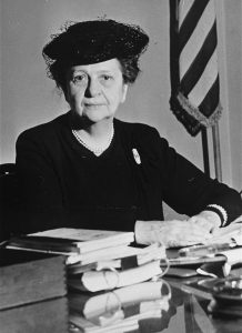 A black and white photograph of Secretary of Labor Frances Perkins in older age, wearing a black beret, at a desk with books.