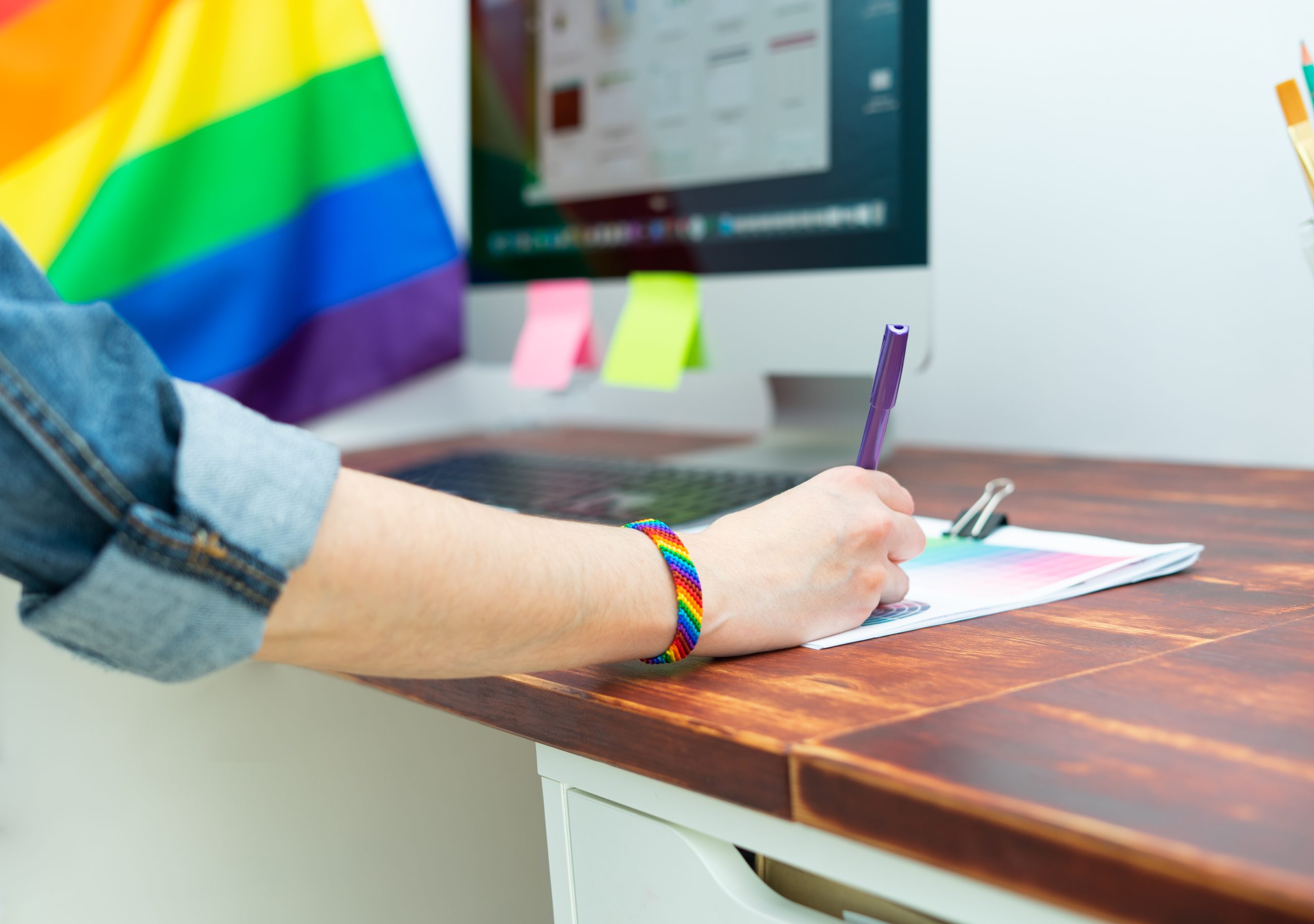 Woman's hand working in office with LGBT decor and accessories. Cultura LGBTQIA LGBTQIA+ inclusive workspace with a person writing wearing a rainbow bracelet, LGBTQIA+ pride flag in background
