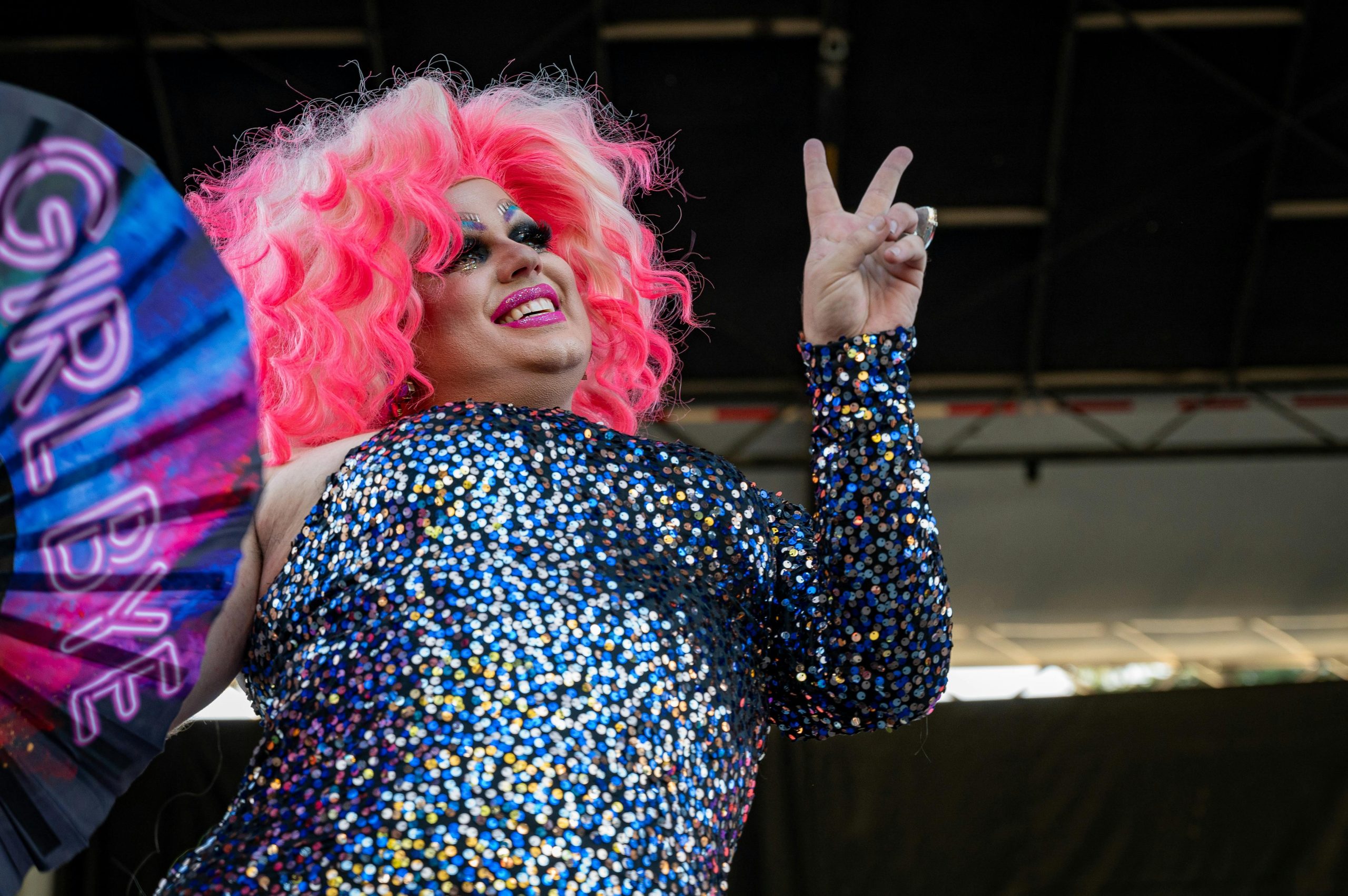 A drag queen with a hot big wig and sequin dress poses for the camera, fingers in a peace sign
