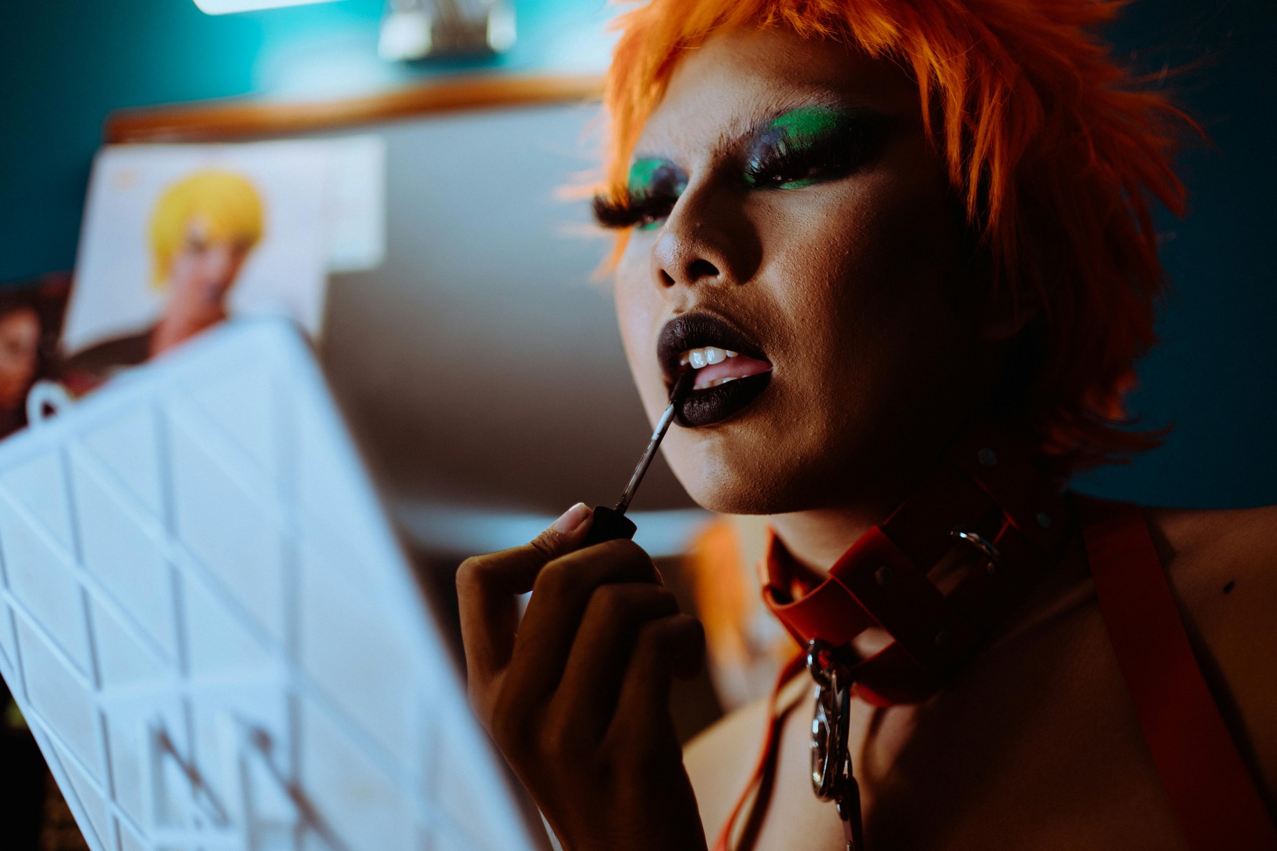 A drag performer applies black lipstick in a handheld mirror. They have bright orange hair and bold green eyeshadow.