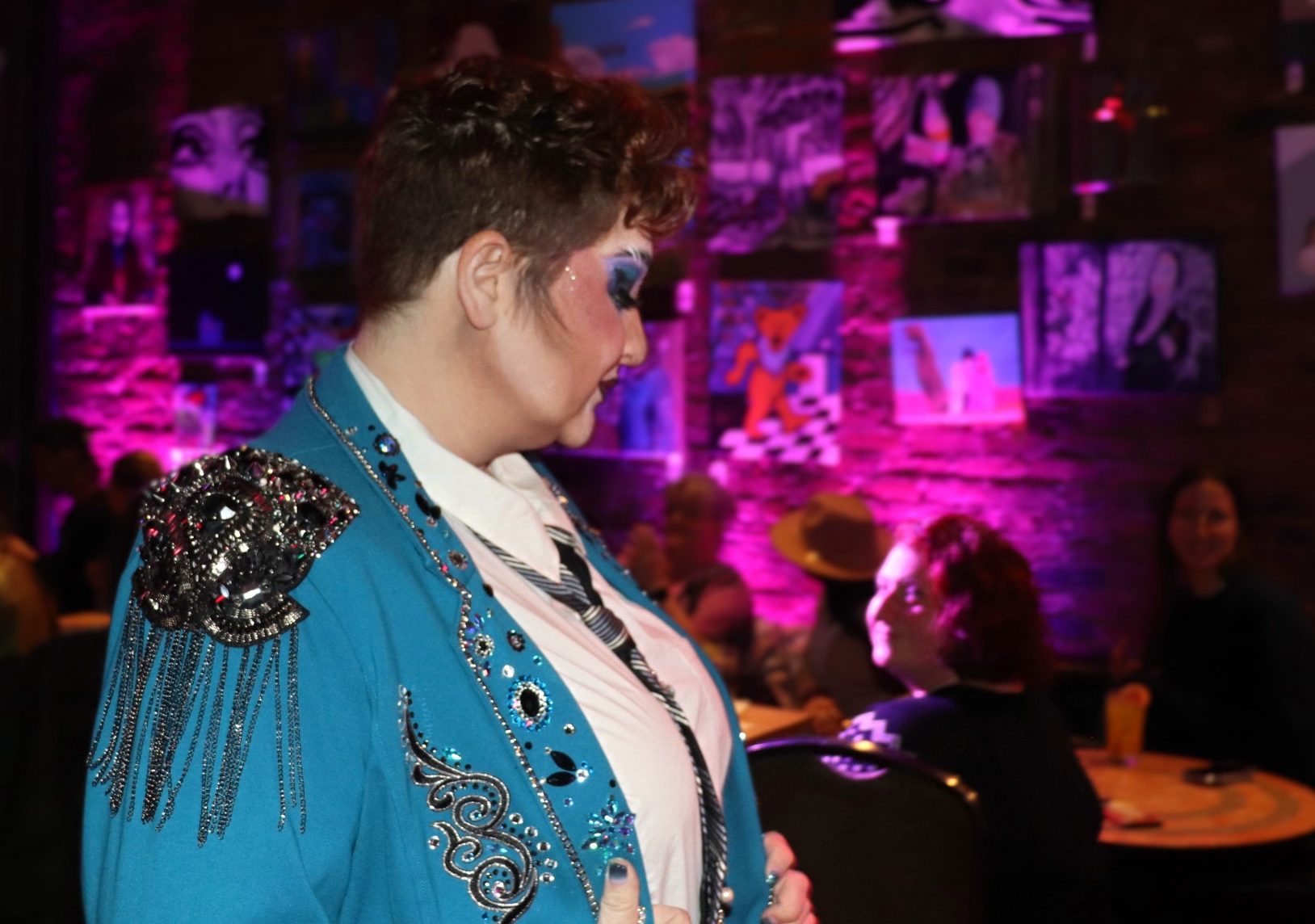 Ali Kane, dressed in drag, looks away from the camera. They have short, curly auburn hair with bold turquoise and black eye makeup. They are wearing a turquoise blazer with black sparkly embellishments.