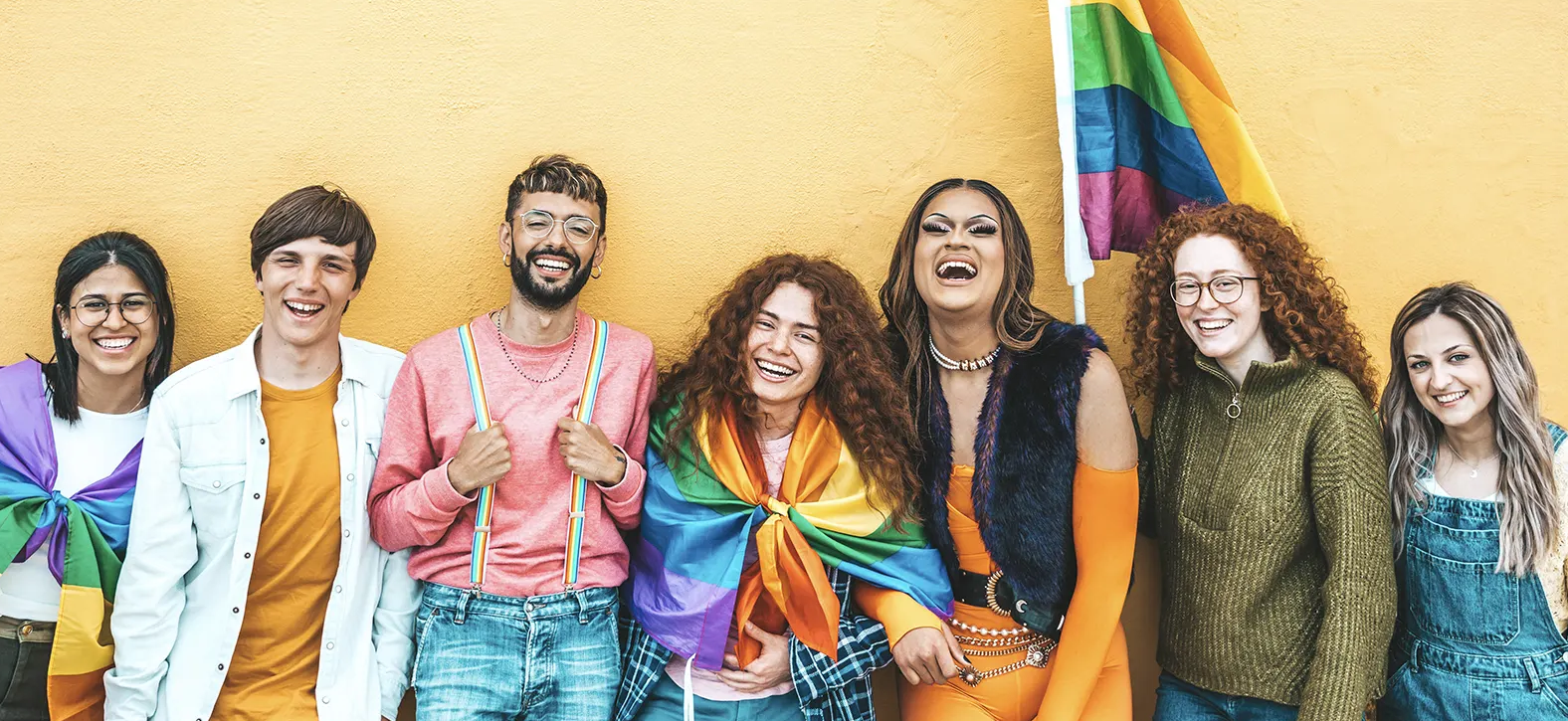 Seven young queer people smile and laugh in front of a yellow background. Several are holding or wrapped in rainbow flags.