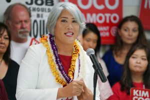 A photograph of Kim Coco Iwamoto at a political rally. She is Japanese Hawaiian woman with short gray hair, wearing a white caridgan and golden lei.