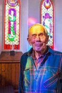 A photograph of James Willis Toy in an church. He is an older Chinese American man wearing a denim shirt.