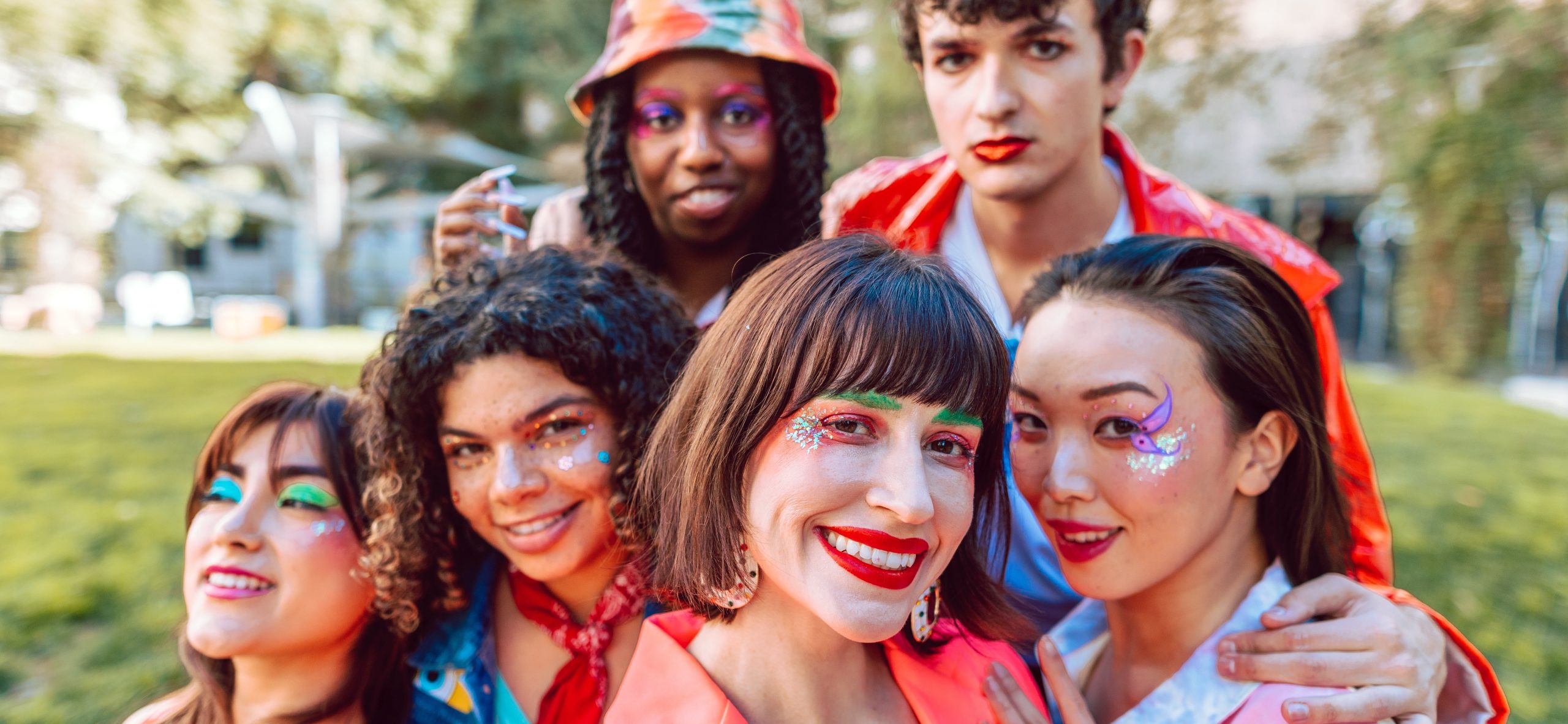 Six queer individuals of varying genders and races, smile at the camera. They all have festive glitter and facepaint.