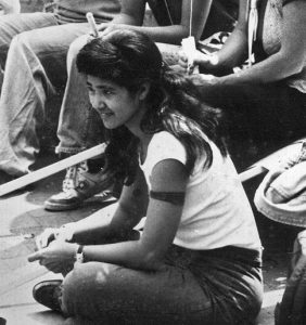 A black and white photograph of Michiyo Fukaya attending the first lesbian gay pride parade and celebration in Burlington, Vermont. She is seated cross-legged on the steps of city hall amount other organizers. 