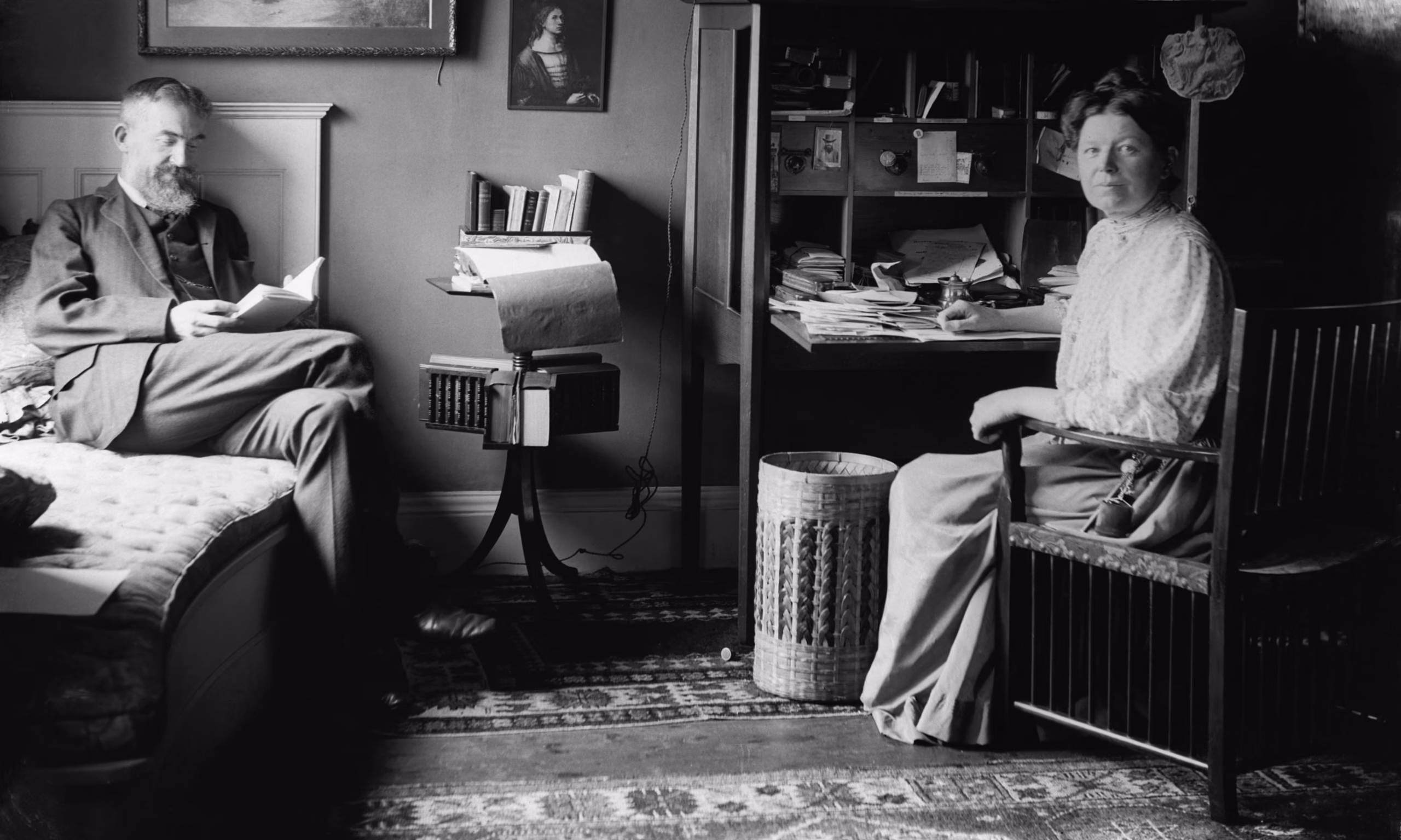 Black and white photograph of Charlotte Payne-Townshend, a middle-aged white woman sitt at her desk. Her husband, poet George Bernard Shaw is read in the corer near her.