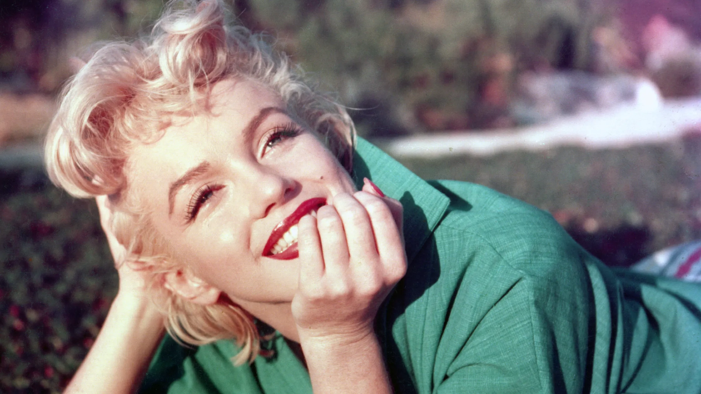 A color image of Marilyn Monroe, styled casually in a jade green collared shirt, resting her head on her hand, and smiling at something off-camera