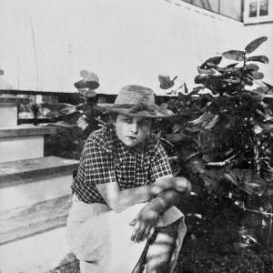 A black and white photo of lesbian poet Elizabeth Bishop, a middle-aged white woman wearing a checkered shirt, light pants, and straw hat, seated on the steps of a home.